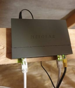 A Netgear network switch installed on a piece of wood in a loft, with three ethernet cables going in and one power cable (for the switch) in the back.